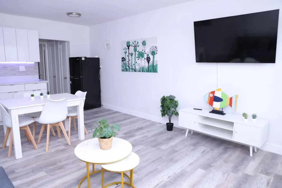 The image showcases a bright, cheerful open-plan living and dining area with a wall-mounted flat screen smart TV, a modern white dining table with stylish chairs, a sleek black refrigerator in the kitchen corner, and tasteful decorative touches like plants and a colorful fish sculpture. The space exudes comfort and modernity, inviting guests to relax, dine, and unwind together.