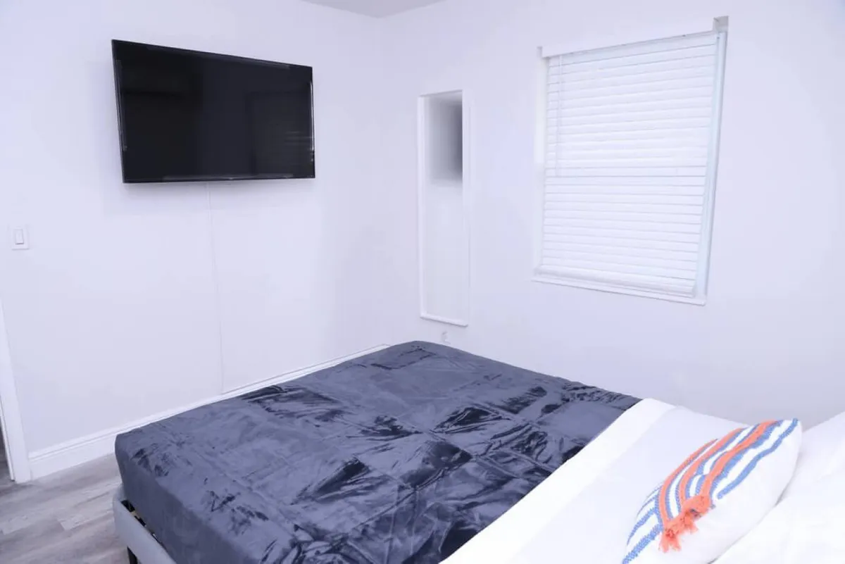 A restful bedroom with a queen bed dressed in a dark blue blanket, a wall-mounted flat screen TV, a window with white blinds, and a small wall mirror. The space is calm and inviting, ideal for relaxing after outdoor activities.