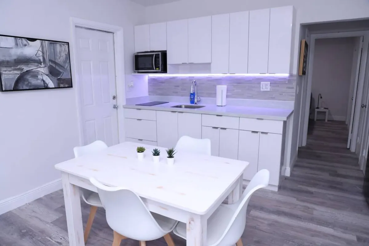 A bright, modern kitchen with white cabinetry and under-cabinet lighting, seamlessly flowing into a dining area with a simple white table and four chairs. Small potted plants add a fresh touch to the table, and a hallway leads to other rooms, giving a sense of openness and connection.