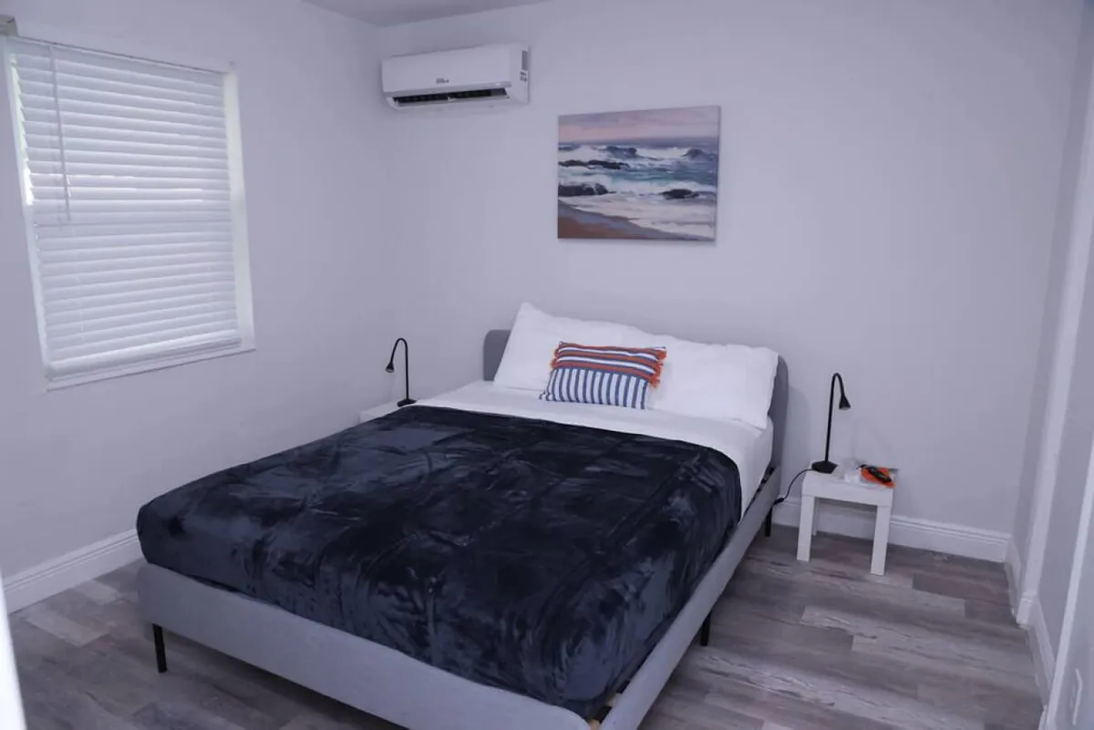A softly lit bedroom featuring a queen bed with a navy blue blanket and striped pillows. The space includes a window with white blinds, a small white bedside table with a reading lamp, wall-mounted air conditioner, and ocean-themed wall art. The ambiance is calm and uncluttered, suggesting a restful retreat.