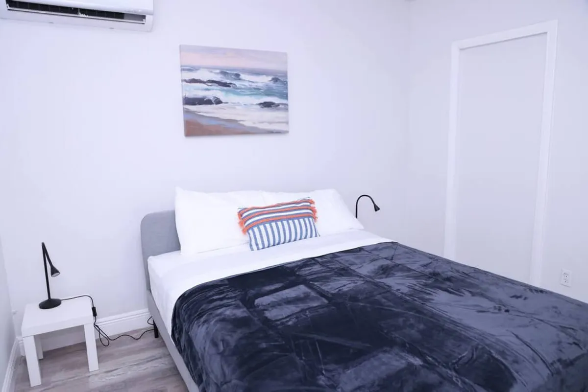 A bright, cozy bedroom centered around a queen size bed with a dark blanket and a colorful striped pillow. Above the bed, a seascape painting enhances the tranquil mood. Minimalist bedside lamps on either side provide soft illumination, inviting relaxation at any time of day.