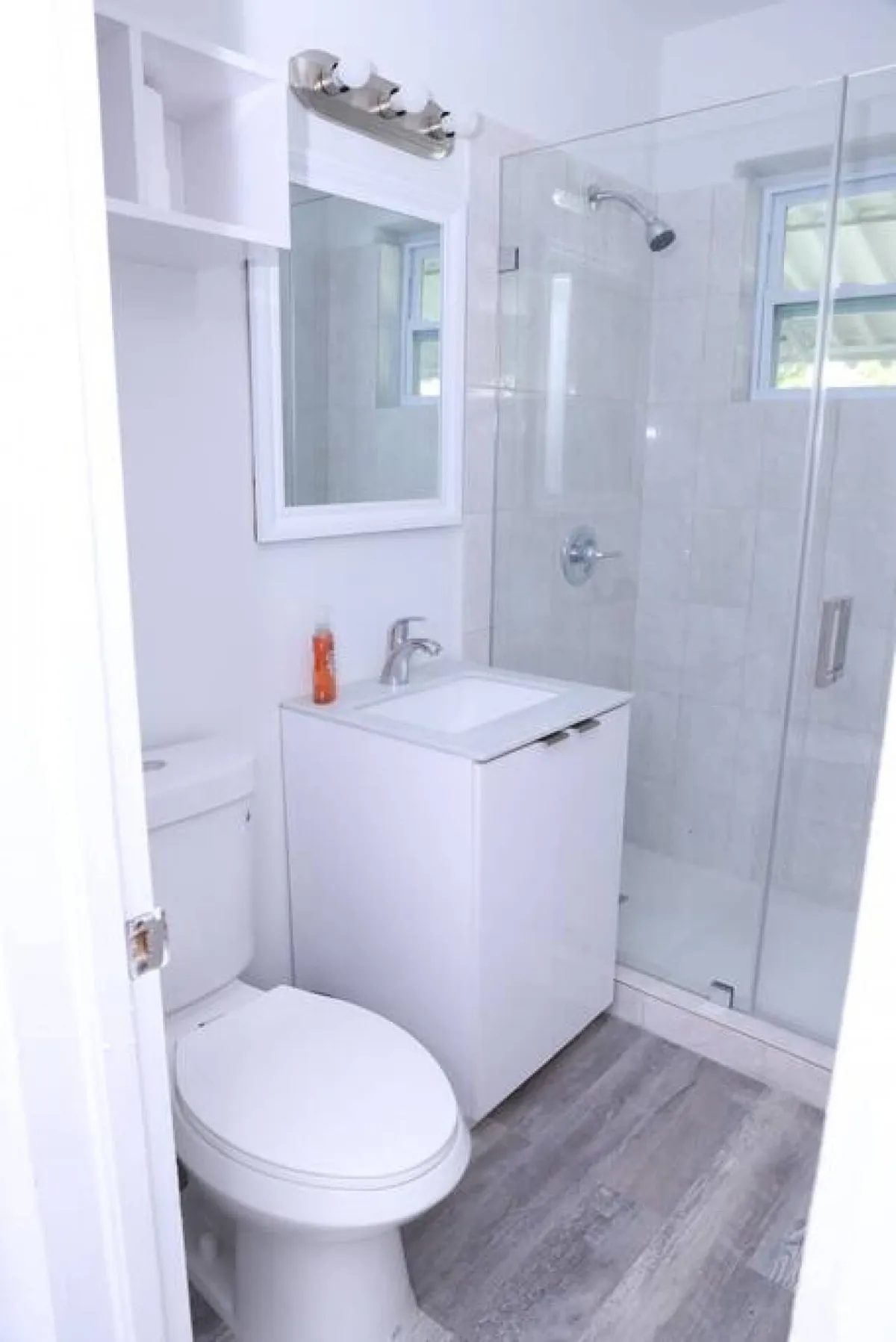 A bright, clean bathroom with a glass-enclosed shower, white vanity with sink, toilet, and a wall-mounted mirror with a light fixture. The space is simple yet functional, emphasizing convenience and comfort.
