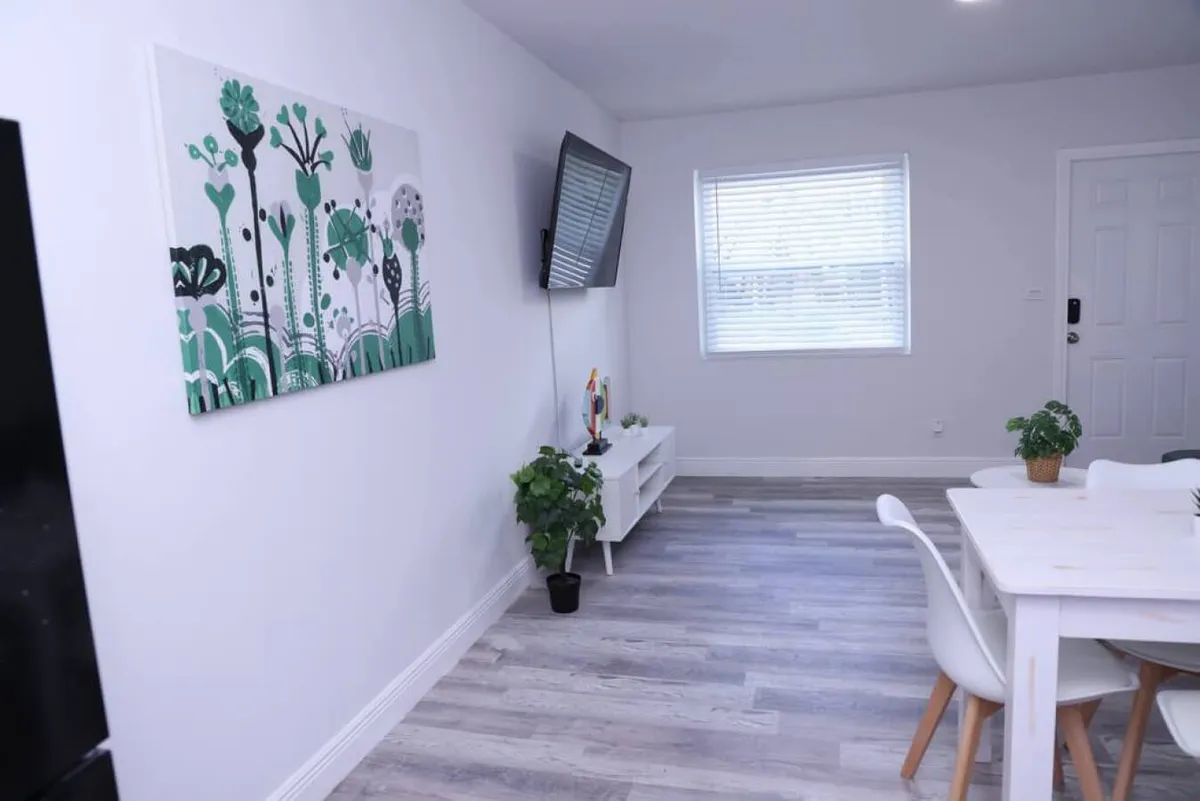 The image shows a cozy living area with a wall-mounted smart TV, a white dining table with chairs, lush green plant decor, and modern abstract botanical wall art. The setting feels fresh and welcoming, with a bright, cheerful ambiance ideal for relaxing or catching up on work.