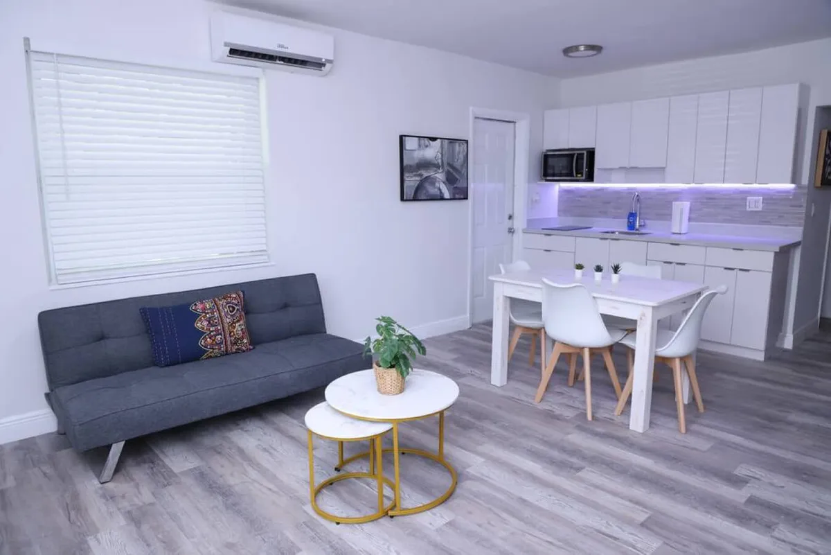 This image features a bright, open-plan living area with a gray sofa bed adorned with decorative pillows, a white dining table surrounded by four chairs, a modern kitchen with under-cabinet lighting and appliances, and round nested coffee tables with a plant centerpiece. The space is airy and functional, with ample light and a layout that encourages relaxation, socializing, and multitasking.