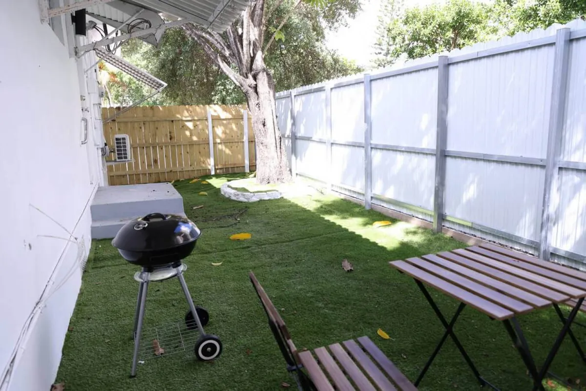 A private patio area featuring a BBQ grill, a set of table and chairs perfect for dining or relaxing, surrounded by a fenced yard with a leafy tree offering shade and privacy. The scene is inviting for outdoor meals, gatherings, or quiet moments with a pet.