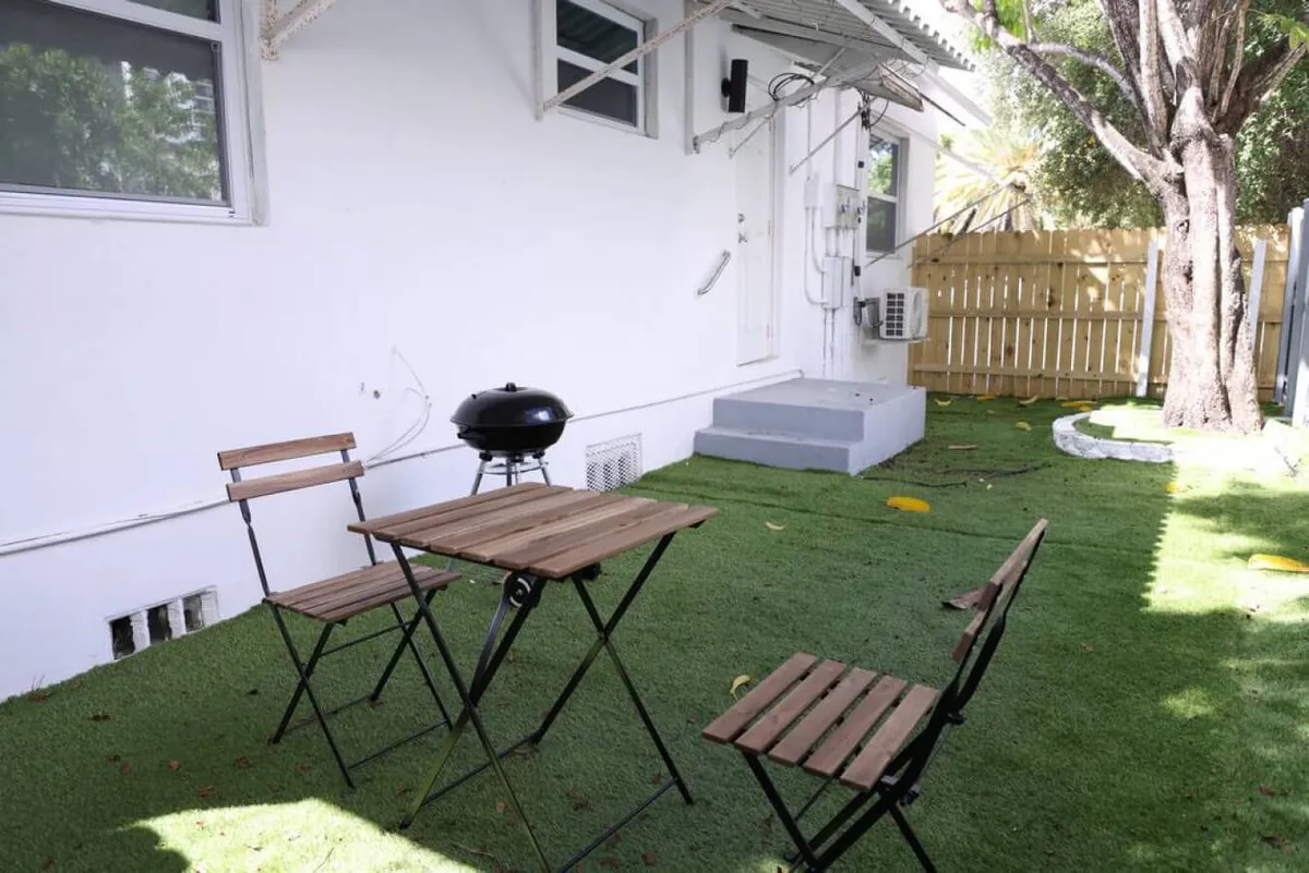 A cozy patio setup with a wooden table, two chairs, a charcoal grill, and a fenced garden with a small lawn area, ideal for outdoor dining, barbecuing, and unwinding. The intimate environment encourages both socializing and relaxation.