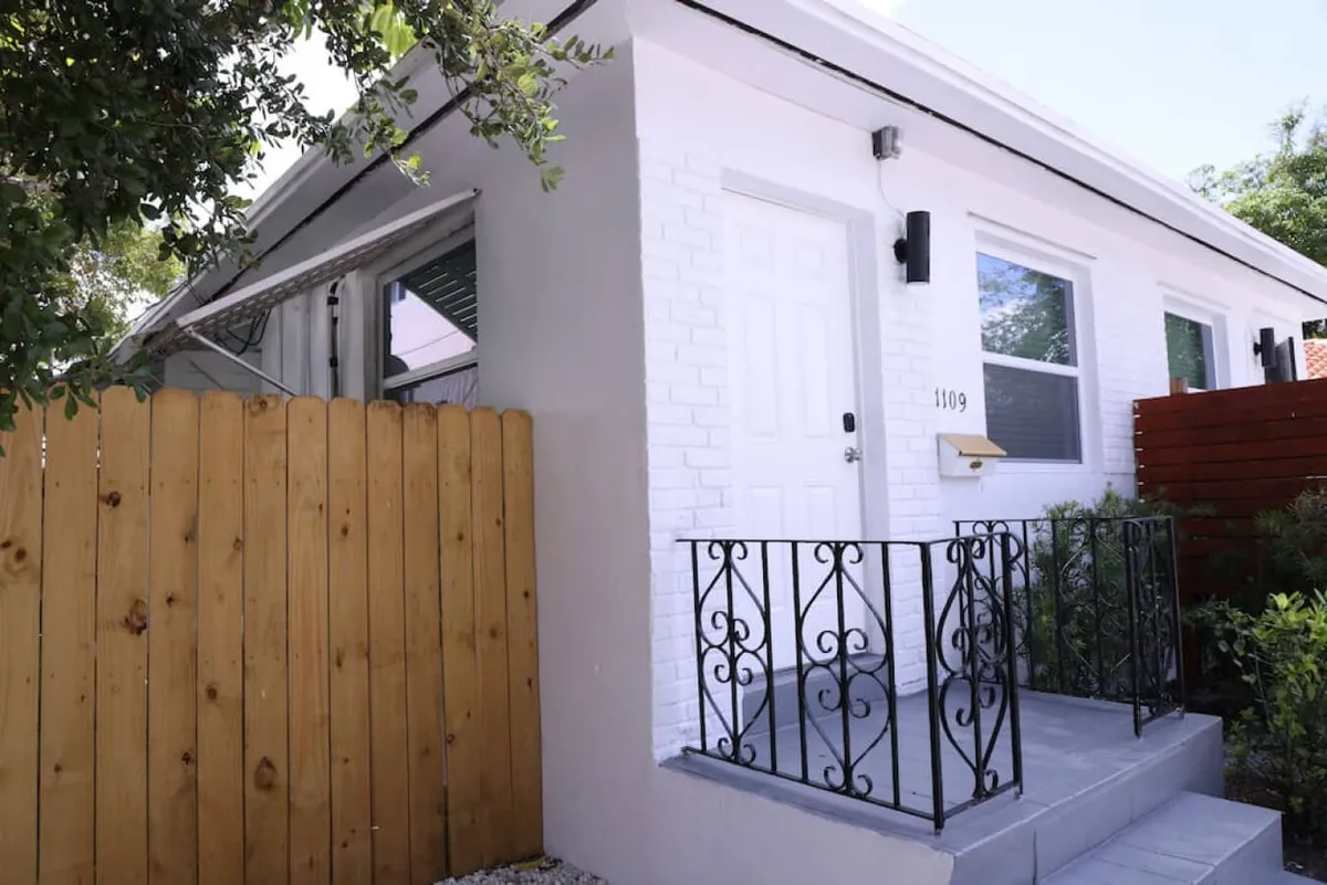 This image displays the inviting exterior of the property, featuring a white brick facade, black wrought iron railing, wooden fence, and lush landscaped plants. The porch and garden areas suggest a welcoming, vibrant atmosphere perfect for relaxing, socializing, or enjoying a sunny afternoon.